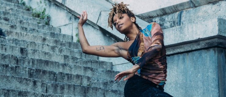a female dancer wearing jeans and a colorful, one-sleeved top posing on stone stairs