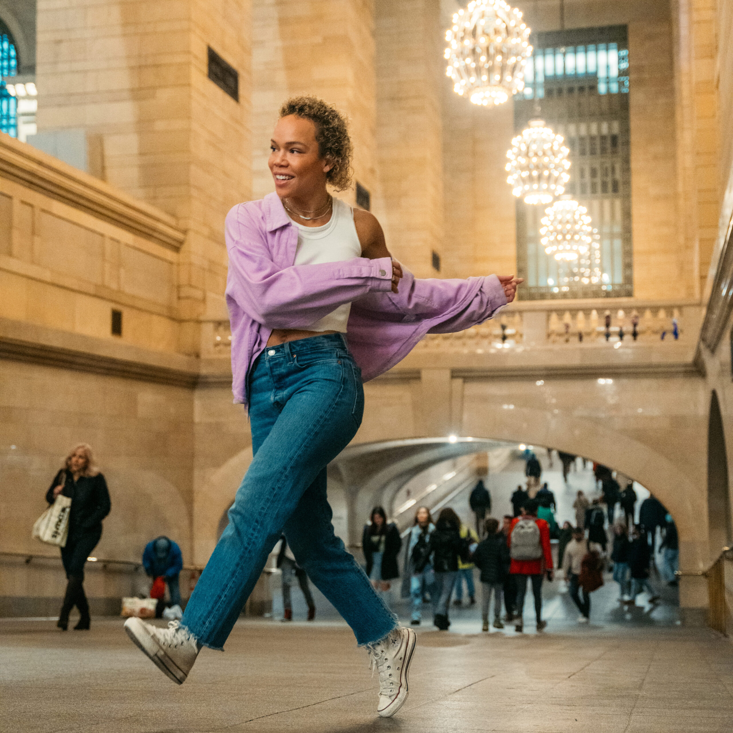 a female dancer wearing jeans, a tank top, and purple button down shirt dancing in a large room with many people walking behind her