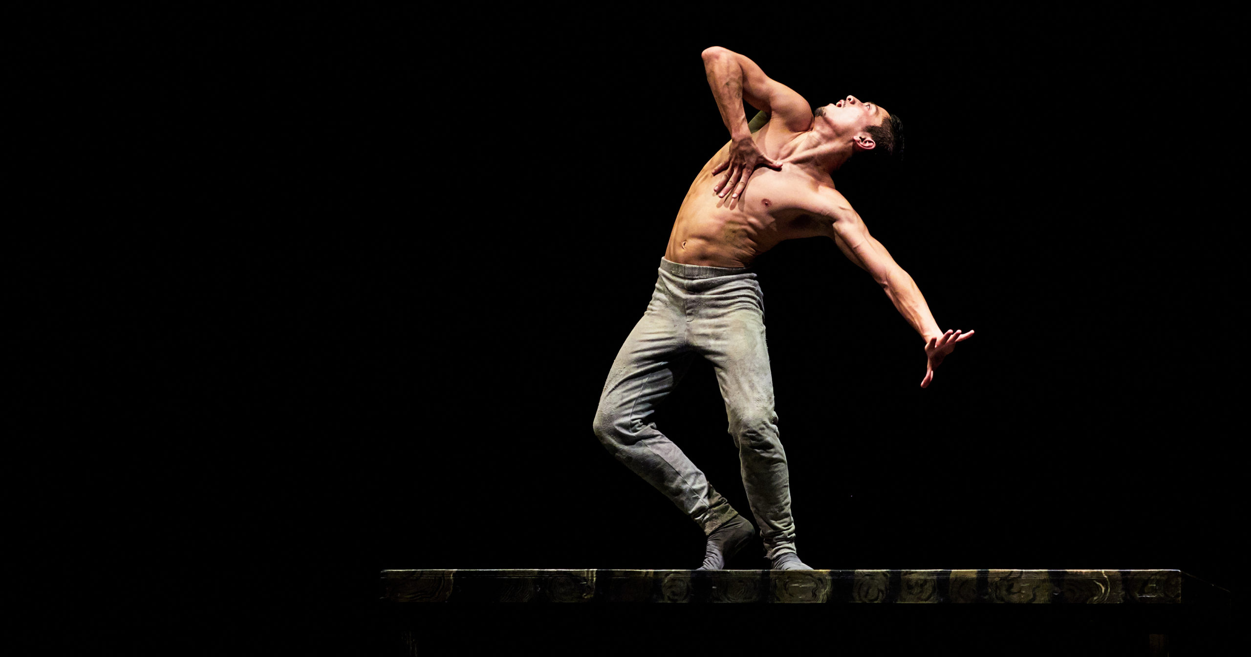 On a darkened stage, Jeffrey Cirio arches back to gaze at the ceiling, one splayed hand over his heart, the other tense to the side. His knees seem to buckle, as though he might sink to his knees at any moment. He is bare-chested and wears tight grey pants.