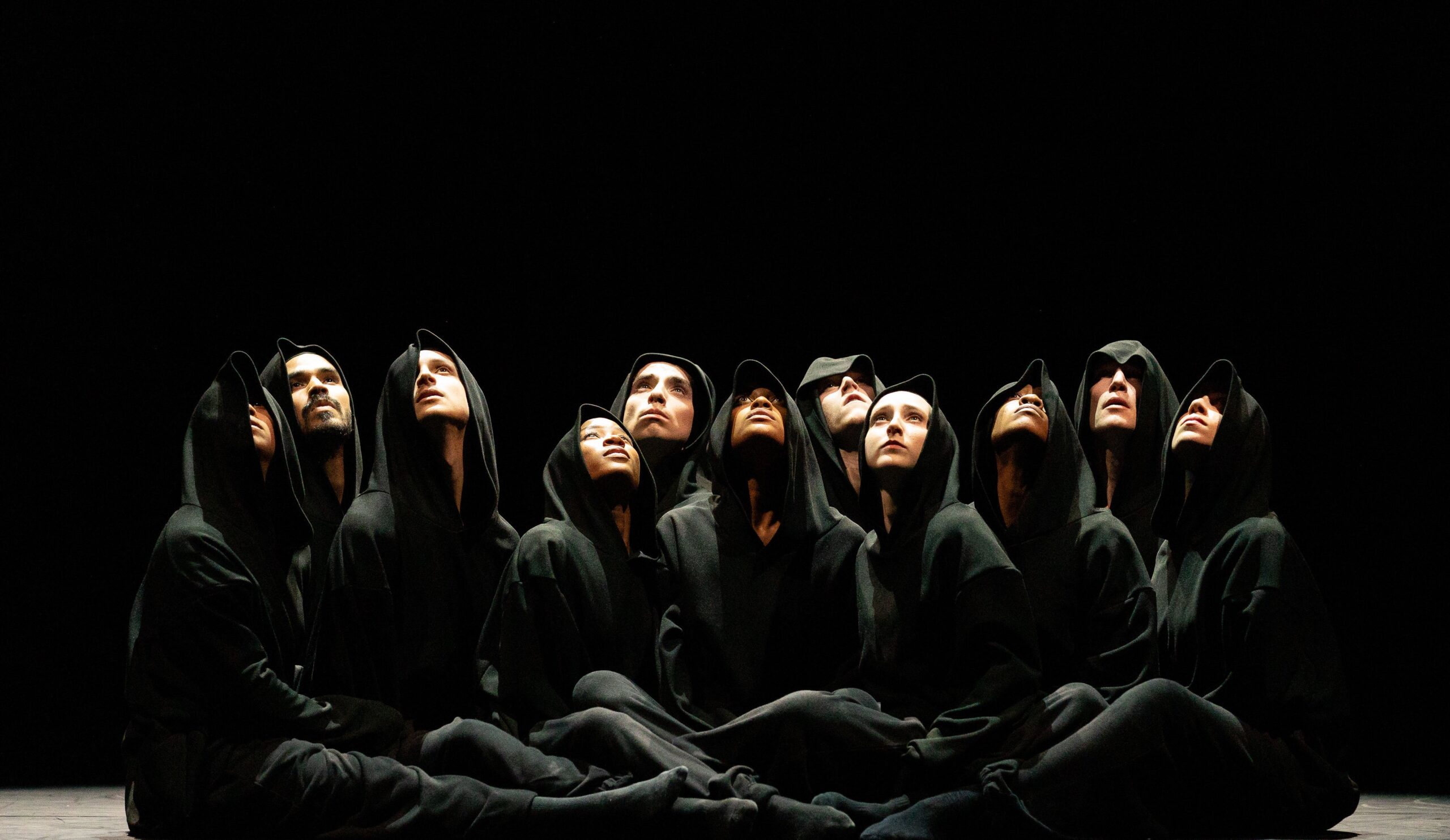 Eleven dancers sit clustered tightly together, their upturned faces peering out from beneath obscuring black hoods.