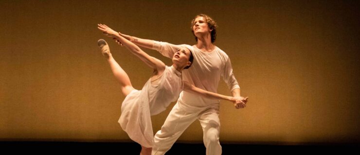 A female dancer moves through a back attitude in parallel, upper body twisting toward her working leg. Her outstretched arms are held by a partner who lunges beside and behind her, gazing in the same upward direction as her.