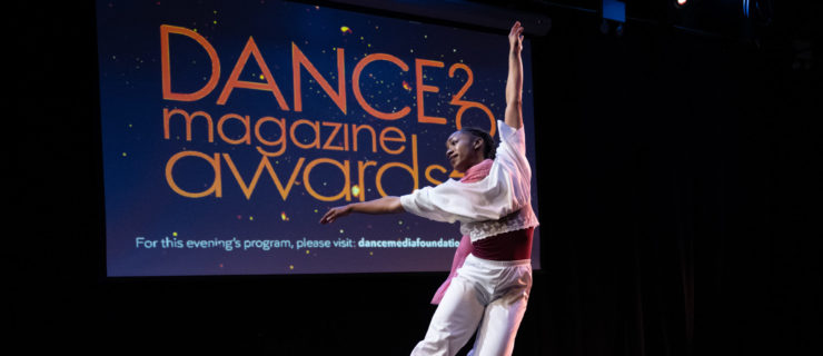 A Black dancer in flowing white pants and a half-sleeved crop top is caught midair in a petite jeté, looking over her side-extended arm in profile. In the background, a projection screen shows the logo for the 2022 Dance Magazine Awards.