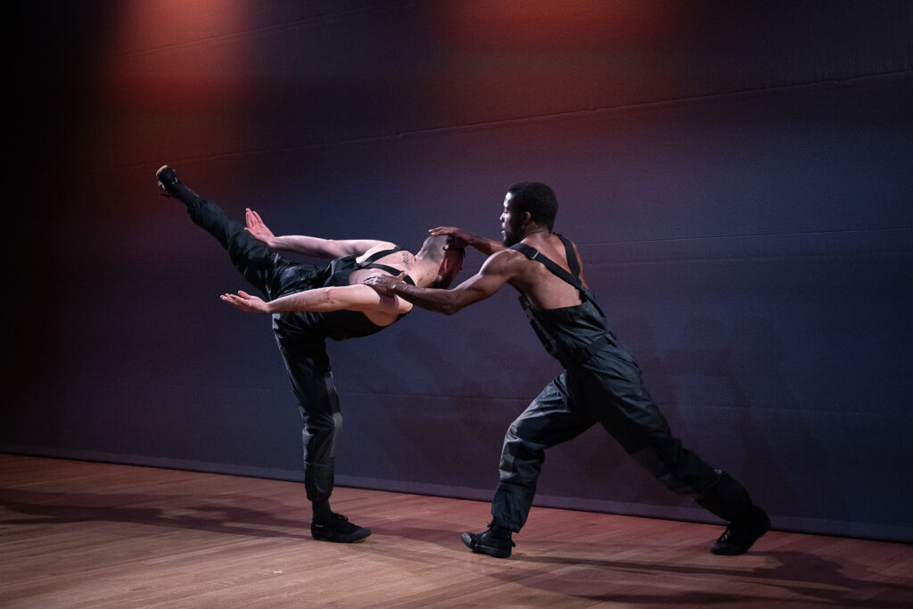 Baye & Asa, two athletic young men, one Black and the other white, in industrial or cyberpunk feeling black overalls. Baye lunges forward to press his hands down on Asa's head and downstage shoulder. Asa's upstage leg swings back in arabesque as his torso flies parallel to the floor. His arms wing back behind him. It feels as though Baye is keeping Asa from flying away.