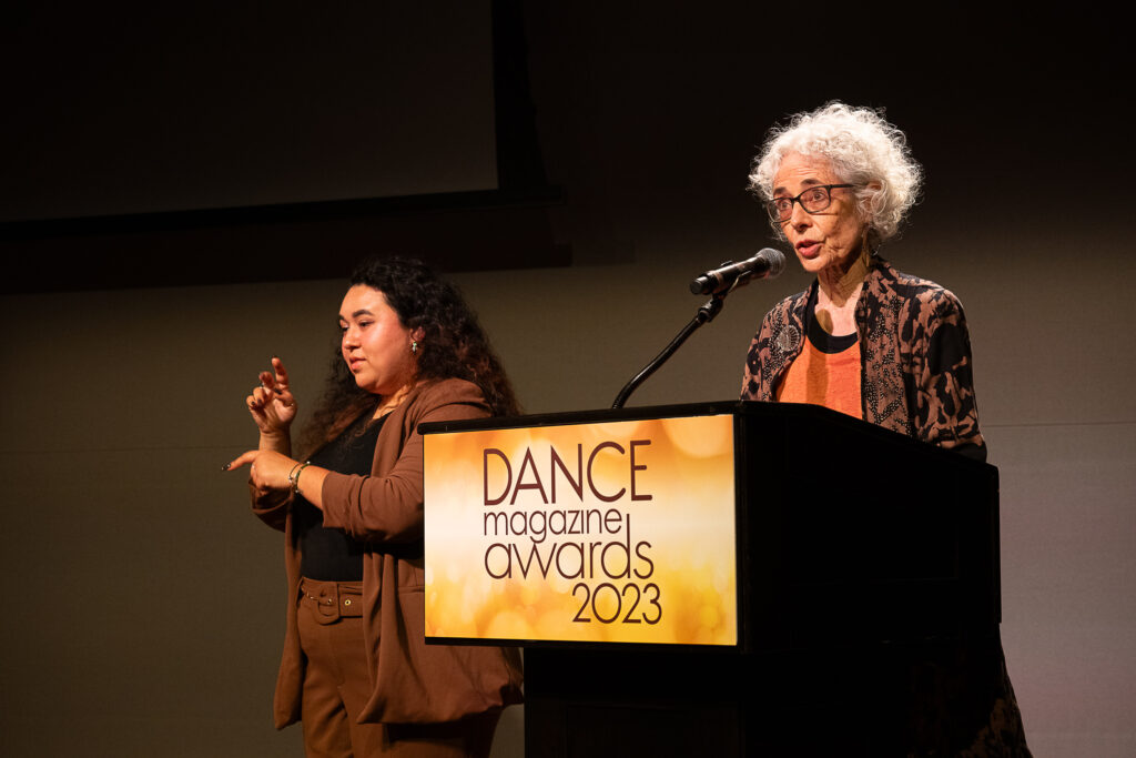 Wendy Perron, an older white woman with fluffy white hair and dark-rimmed glasses, speaks intently into a microphone at a podium with a sign that reads "Dance Magazine Awards 2023." To her right is an ASL interpreter.