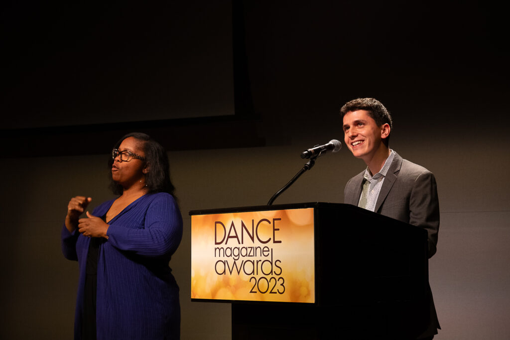 Eric Oberstein, a young white man in a tieless grey suit, smiles as he speaks into a microphone at a podium with a sign that reads "Dance Magazine Awards 2023." An ASL interpreter is at his right.