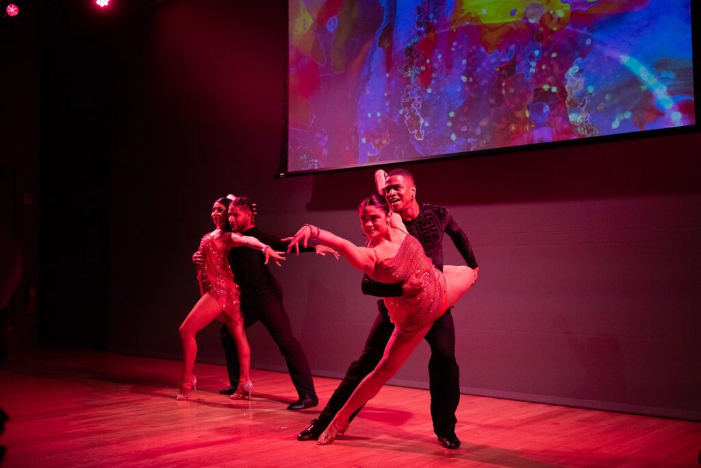 Two couples performing a Latin ballroom routine pose on a stage awash in red light.