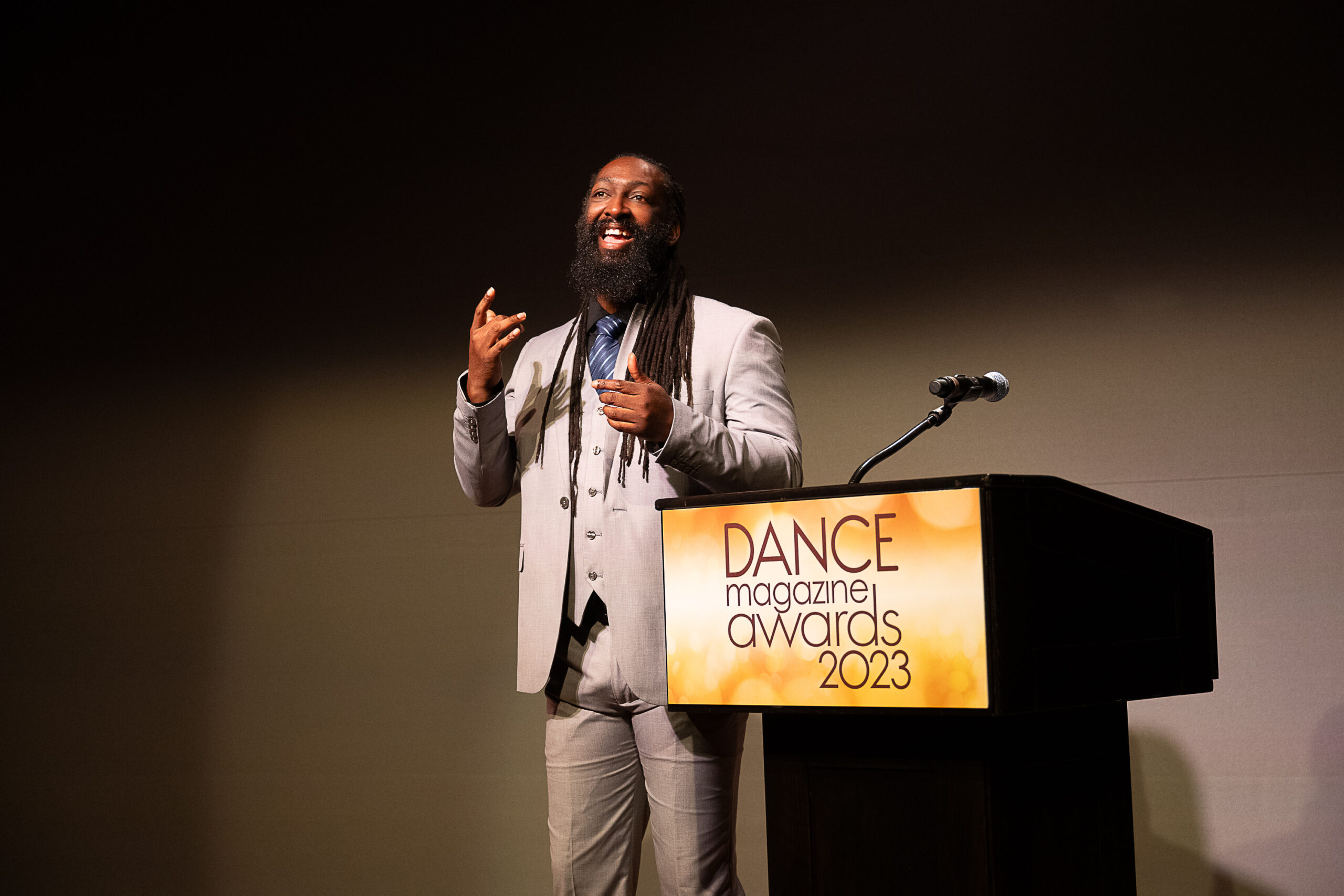Antoine Hunter Purplefirecrow, a Black and Indigenous person with long dreads and a bushy beard. He wears a sharp, pale grey suit. He gives an infectious smile as he signs, standing beside a podium emblazoned with the logo for the 2023 Dance Magazine Awards.
