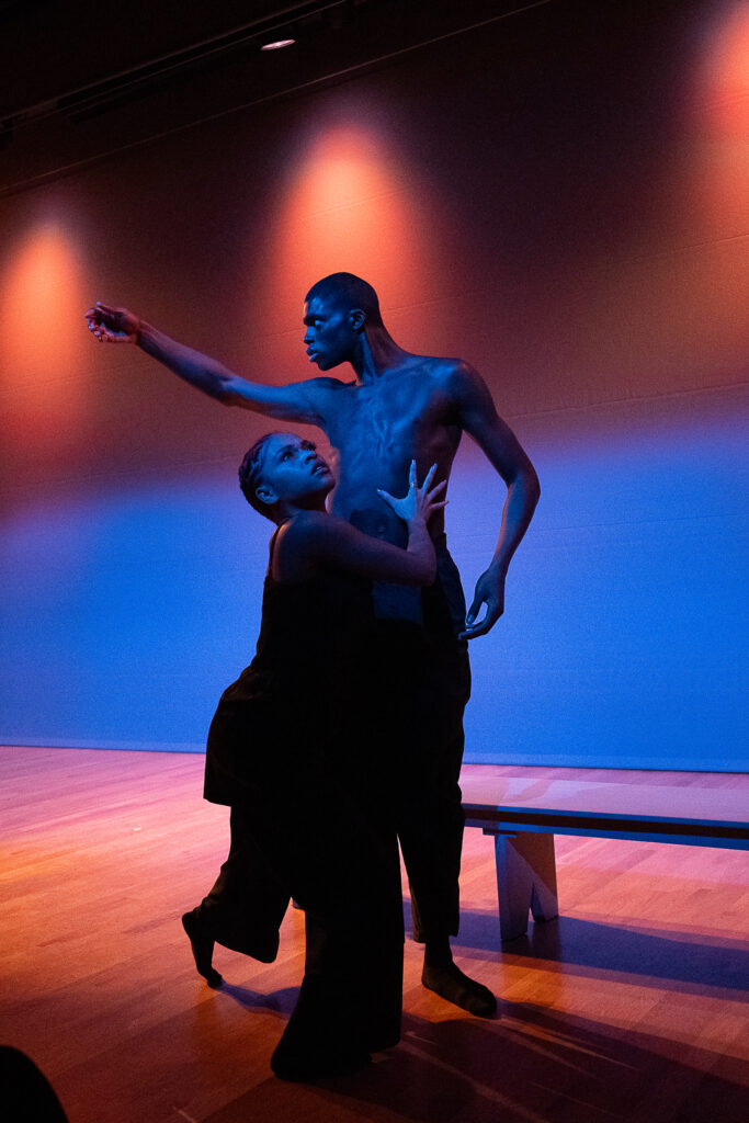 Two young, Black dancers perform on a stage awash in blue and orange light. Waverly Fredericks extends a curving arm to the side, head craning forward to look where it is pointing. Rachel Lockhart steps forward in front of him, her eyes cast upward and away as she presses her palms to his ribcage.