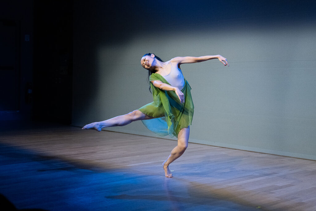 Frances Lorraine Samson, a petite but athletic woman of Asian descent, smiles radiantly as she balances on forced arch. Her arms pull to one side as her leg extends to the other, her head tipping toward her working leg. She wears a costume of flowing green material over a nude leotard; her black hair is pulled back from her face but otherwise allowed to flow freely behind her.