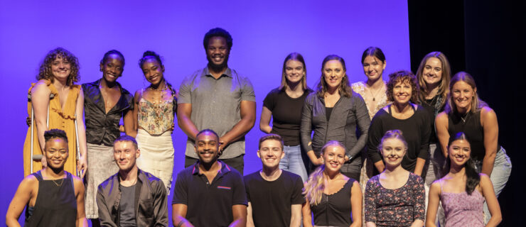 Seventeen of the 2023 25 to Watch Live! performers pose for a group photo on the Sheen Center stage. They stand or kneel and wear nice street clothing.