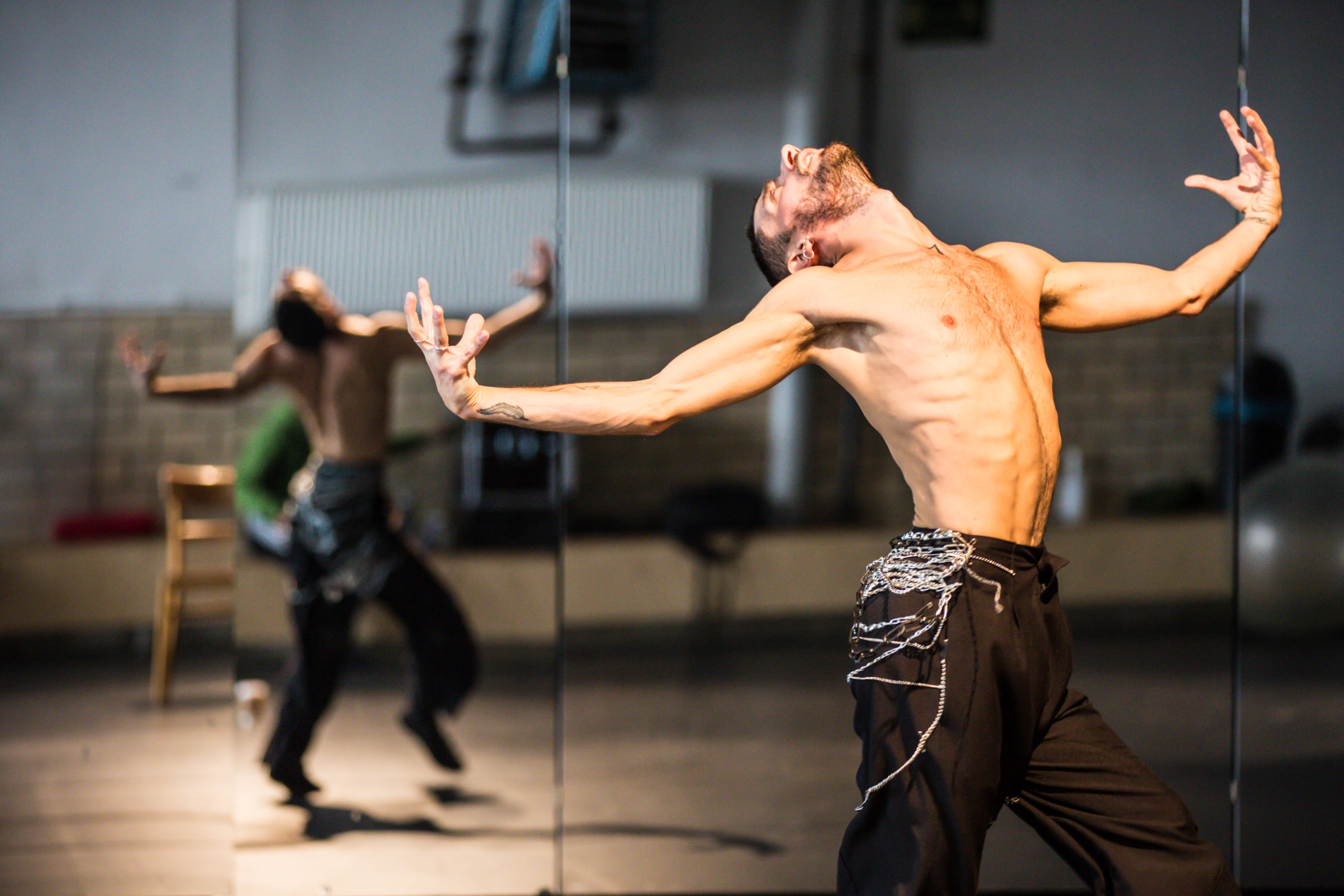 A shirtless male dancer sits into one hip, head arched back to look at the ceiling, his arms curving as they extend to either side with his hands curling into claws. His back is reflected in the studio mirror behind him.