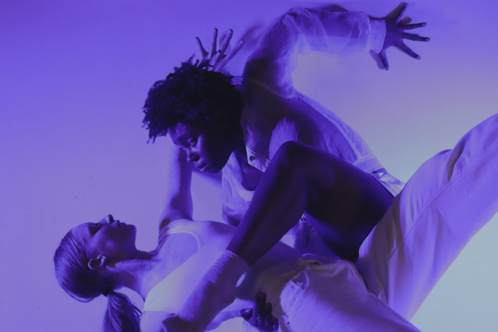 Ashley Green, wearing white and lit purple, gazes intently down at another dancer as she supports her with an arm around her waist. Green's downstage leg crosses her partner's torso in a parallel attitude; her upper arm creates a right angle behind her, palm splayed.