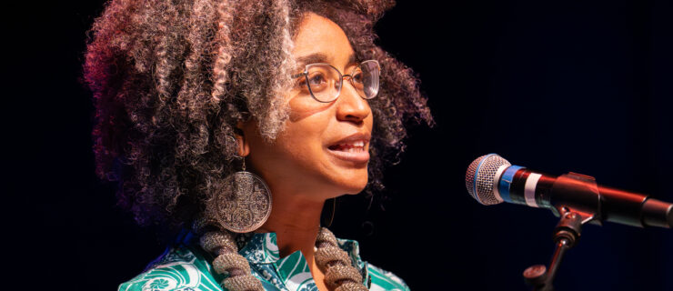 Gumbs stands at a podium, speaking into a microphone, wearing a patterned green shirt and chunky brown jewelry.