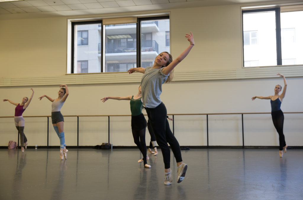 Adriana Pierce demonstrates at the front of a studio for five women on pointe. She moves through fifth position on relevé with her back foot raised, downstage arm overhead, with a slight arch in her upper back. She wears worn white converse, black leggings, and a grey shirt. Her dirty blonde hair is worn loose.