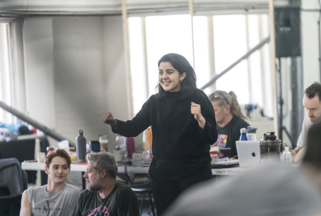 Arielle Smith stands at the front of a studio, smiling encouragingly as she raises both fists to chest height, eyes fixed on the dancers in the space. In the background, individuals sit with laptops and water bottles at a long table.