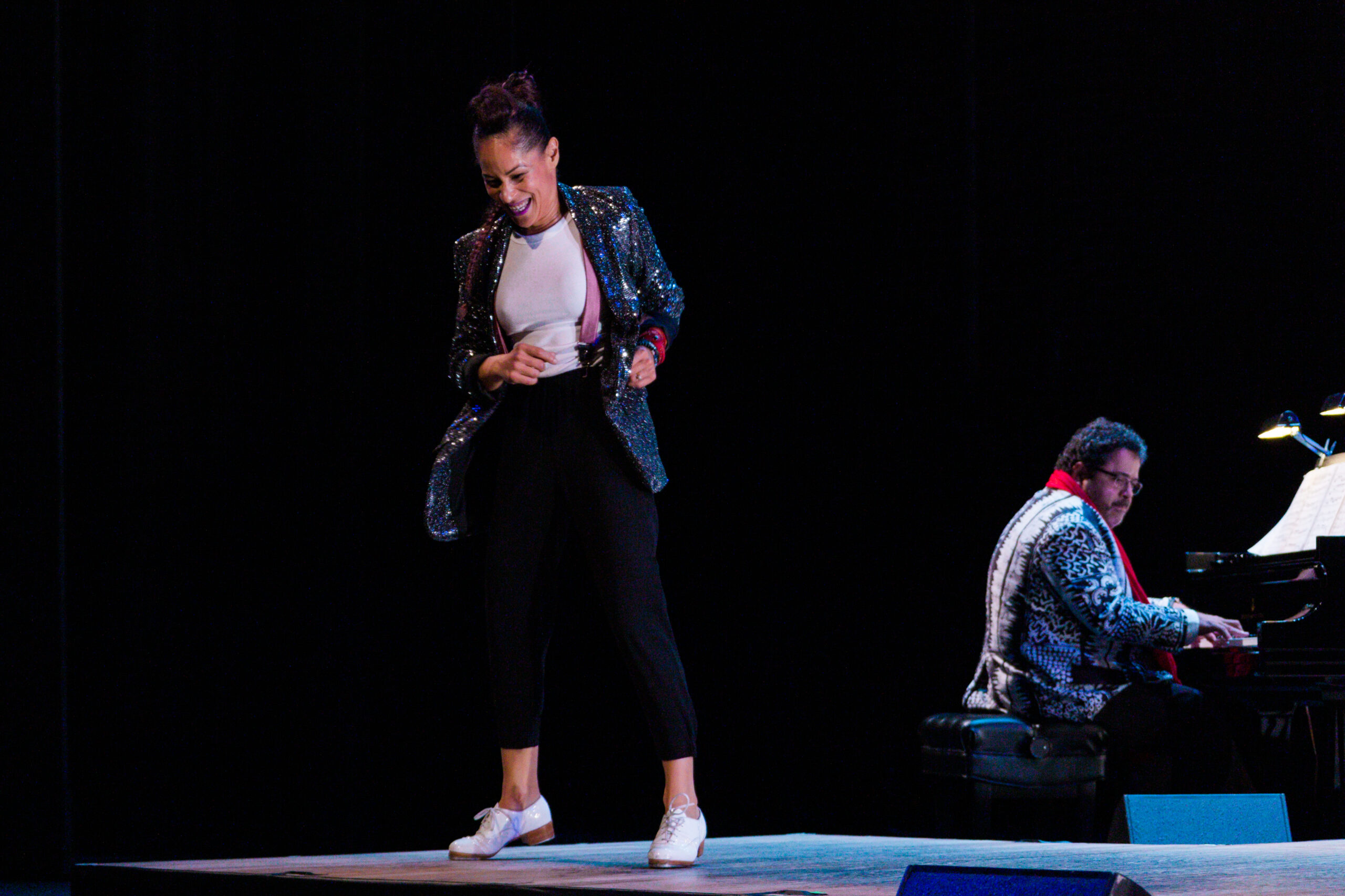 a female tap dancer front and center with a man playing the piano in the right corner