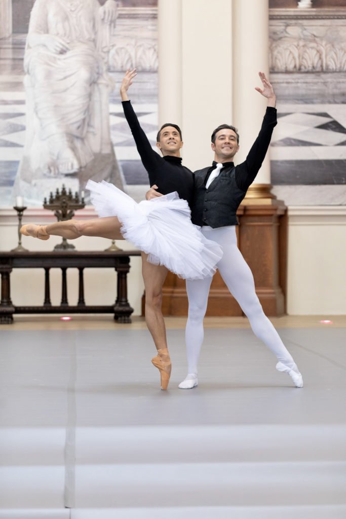 Carlos Hopuy, in pointe shoes, a white classical tutu and black turtleneck, poses in an open attitude back on pointe. Diego Cruz supports him with an arm around his waist, the other mirroring Carlos' high fifth; he wears more traditional white tights. Opulent paintings and classical pillars are visible beyond the grey marley floor.