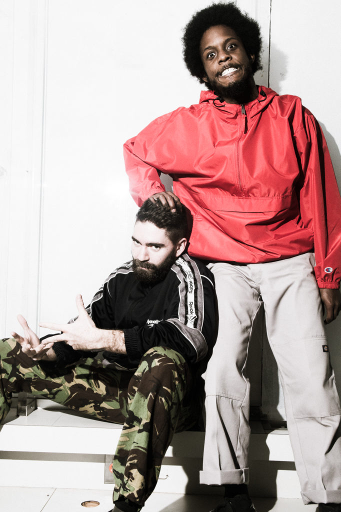 Amadi Baye Washington pulls a wide-eyed, open mouthed face just past the camera as he presses a hand into Sam Asa Pratt's curly hair. Pratt sits on a bench, elbows on his knees and fingers splayed as he looks intensely in the same direction. Pratt wears camo pants and a black sporty long sleeved shirt; Washington wears light grey athletic pants and a bright coral jacket.