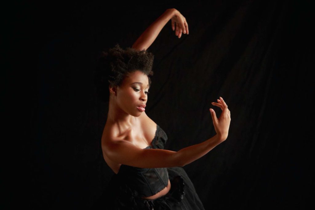 Brianna Mims poses against a black backdrop. Her gaze is cast down towards the graceful curve of her right arm, which she leans away from.