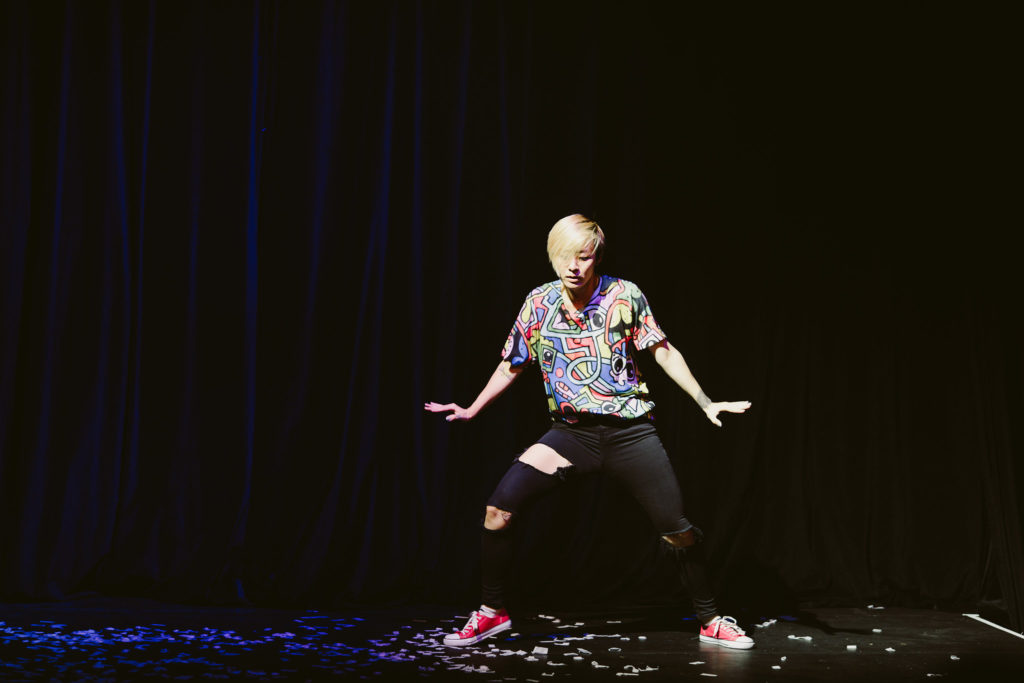On a dark stage, Bo Park moves through a wide stance, arms lightly extended to either side with palms flexed, eyes downcast. She wears red converse, ripped black skinny jeans, and a cartoonish, colorful t-shirt.