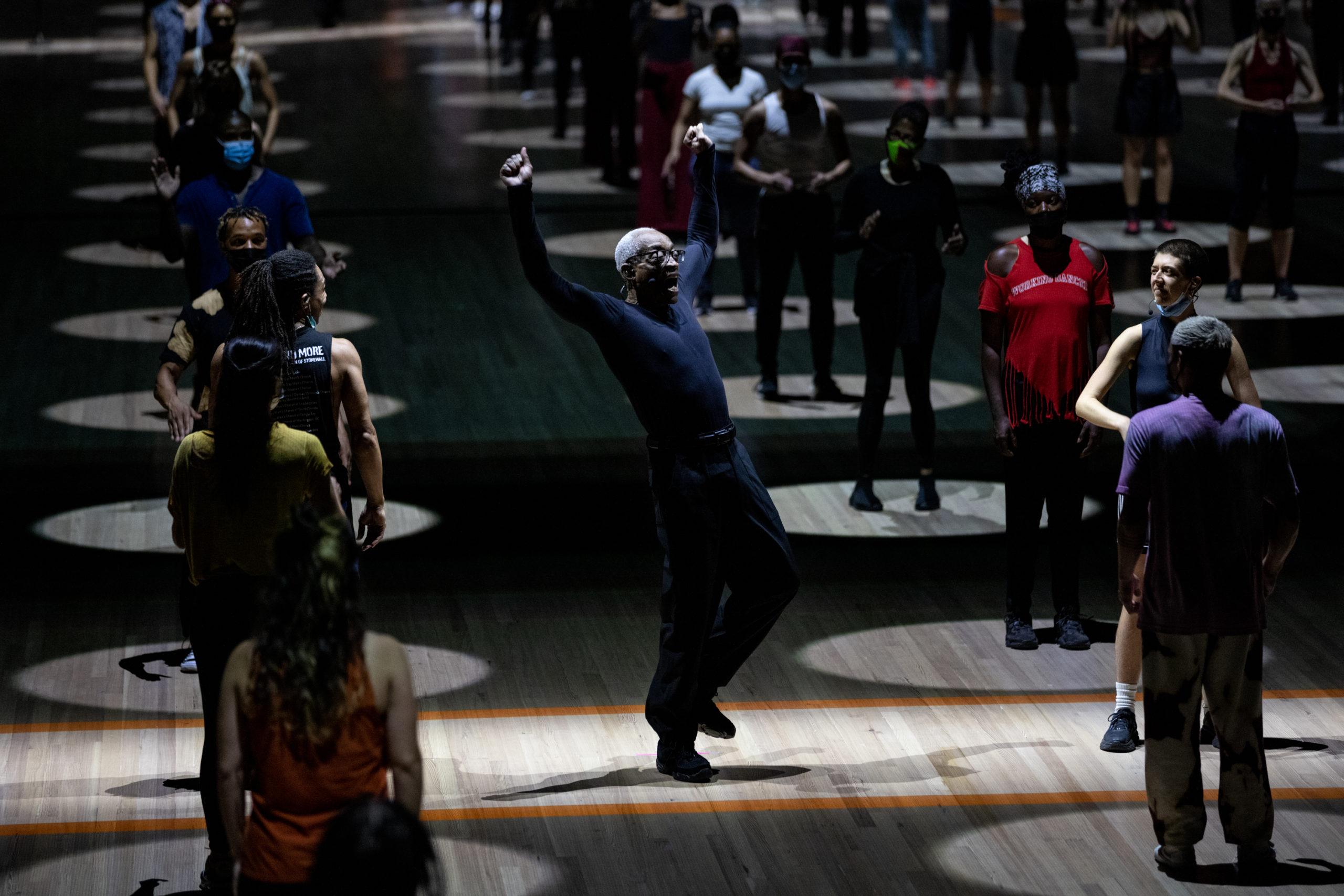 Bill T. Jones, an older Black man, balances with one flexed foot raised slightly off the ground. His arms rise beside his head as his torso leans back. Around him are lines of audience members standing in evenly spaced spotlights.