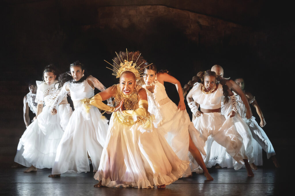 a female dancer wearing a large crown leading a group of dancers in flowy white costumes on stage