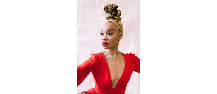 Camille A. Brown looks over her right shoulder. She wears a red blouse with a plunging neckline; her lips are painted the same color. A headscarf with a gold filigree pattern is wrapped around her scalp and some of the hair piled atop her head.