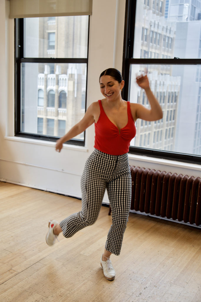 Christina Carminucci, dark hair slicked back from her face, grins and looks down as her white tap shoes blur with motion. She wears black and white check trousers over a red leotard. Tall buildings are visible through the windows behind her in the studio.