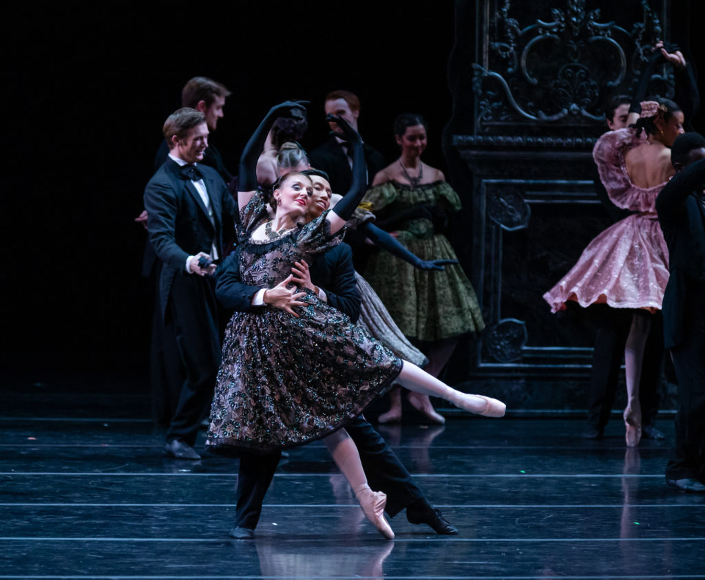 Courtney Nitting wears an opulent dusty purple and black dress over pink tights and pointe shoes, as well as black elbow length gloves. She poses in a low, off-center arabesque, arms in high fifth, supported from behind by a male dancer in an old-fashioned black suit. In the background are similarly costumed dancers.