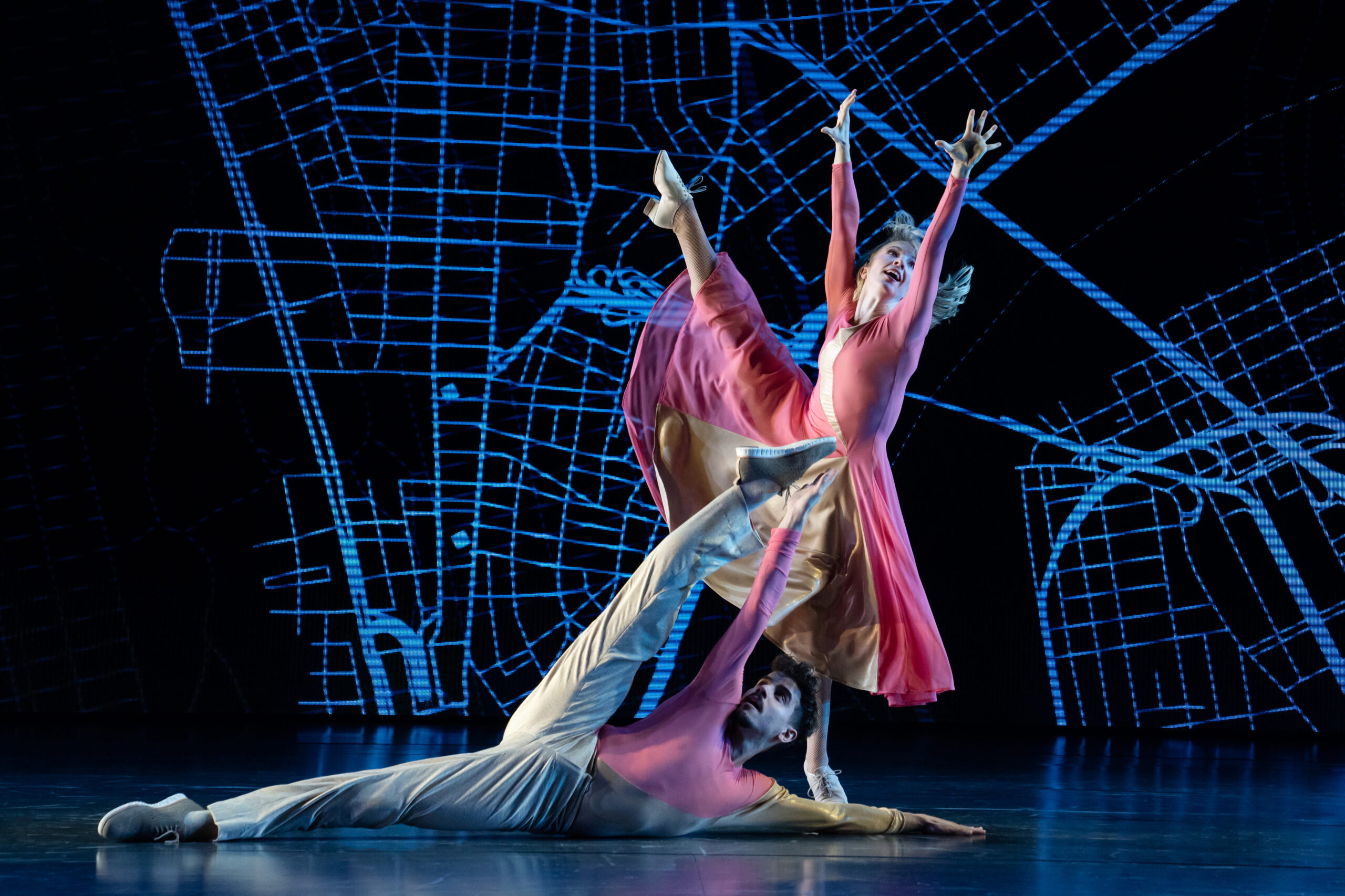 a female dancer reaching up and extending her leg side; a male dancer laying on the floor and kicking over his head
