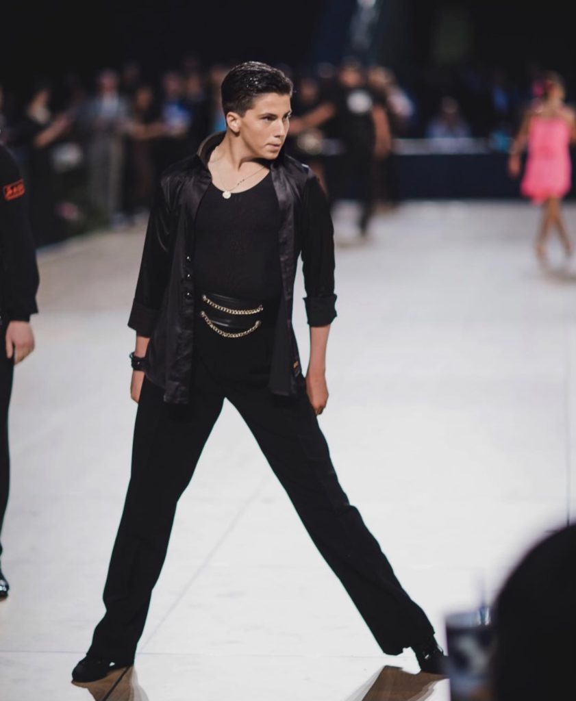 Carter Williams, dressed all in black with a pair of gold chains at his waistcoat, levels an intense look off camera as he strikes a pose in a wide stance, arms by his sides. A crowd and other dancers dressed for ballroom are blurry in the background.