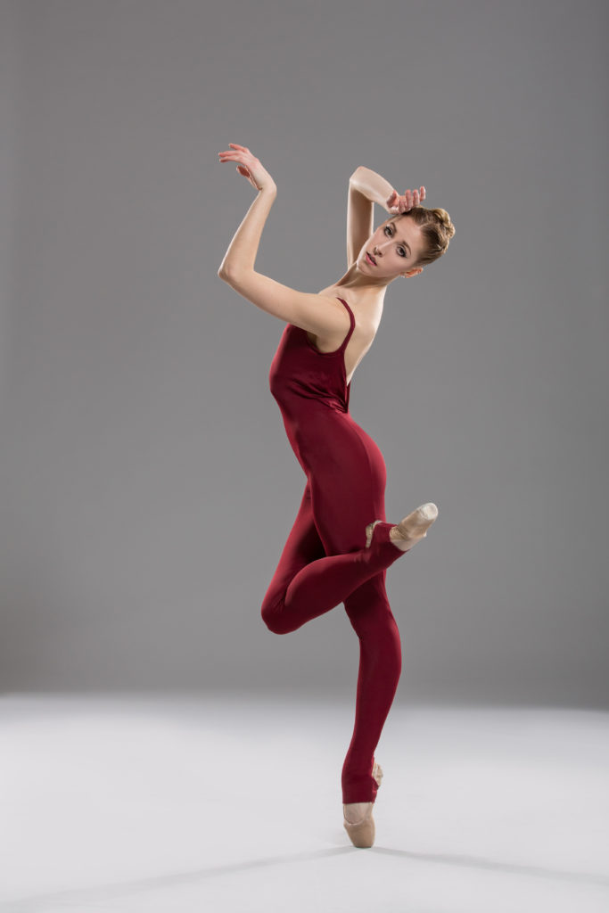 Darian Kane poses in profile in pointe shoes and a red bodysuit against a grey backdrop. She balances on pointe, one knee hooked over the other, arching back slightly as her arms sculpt the air around her face. Her head tips sideways so she can gaze at the camera.