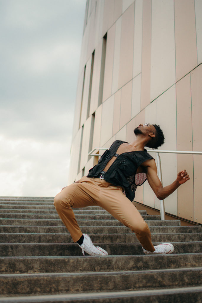 Darvensky Louis gazes upward as he arches back, resisting gravity as he bends over the top of his front foot. He is on an outdoor staircase, wearing white sneakers, brown pants, and a loose black vest that leaves much of his chest exposed.