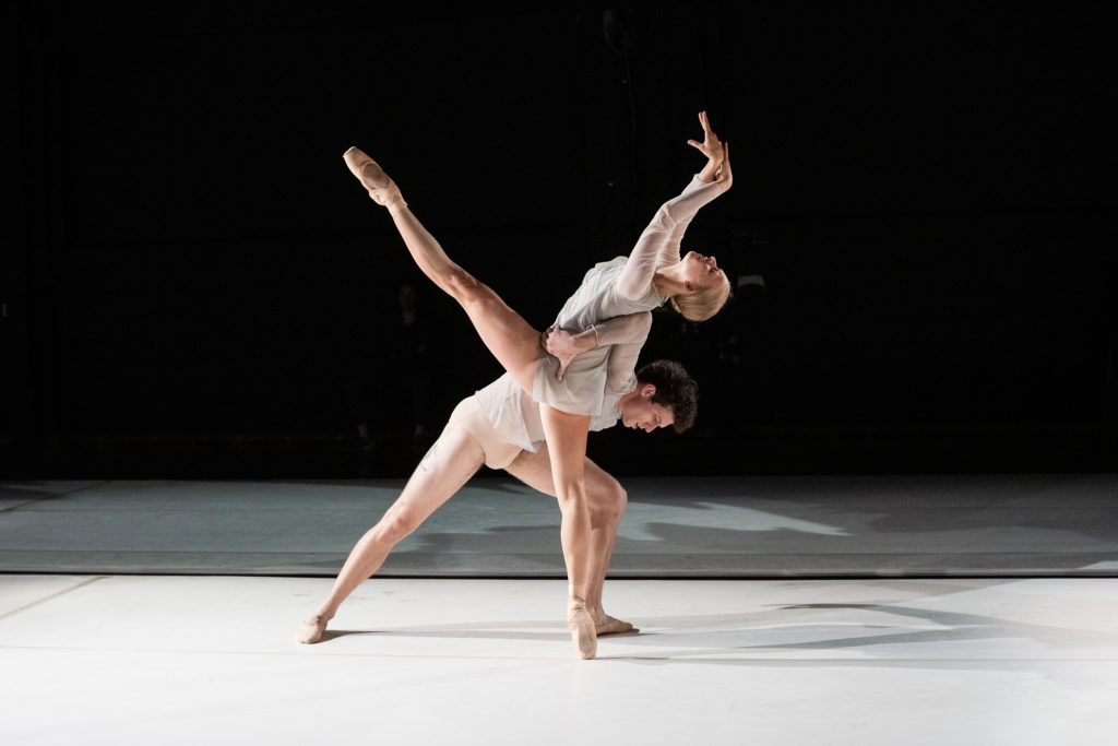 Genevieve Penn Nabity, in pointe shoes, bare legs, and a simple tunic, balances on pointe with one leg extended long in front of her. She arches back, head parallel to the floor and arms extended in front of her. Another dancer lunges beneath her, balancing her with an arm wrapped around her back to her working side hip.