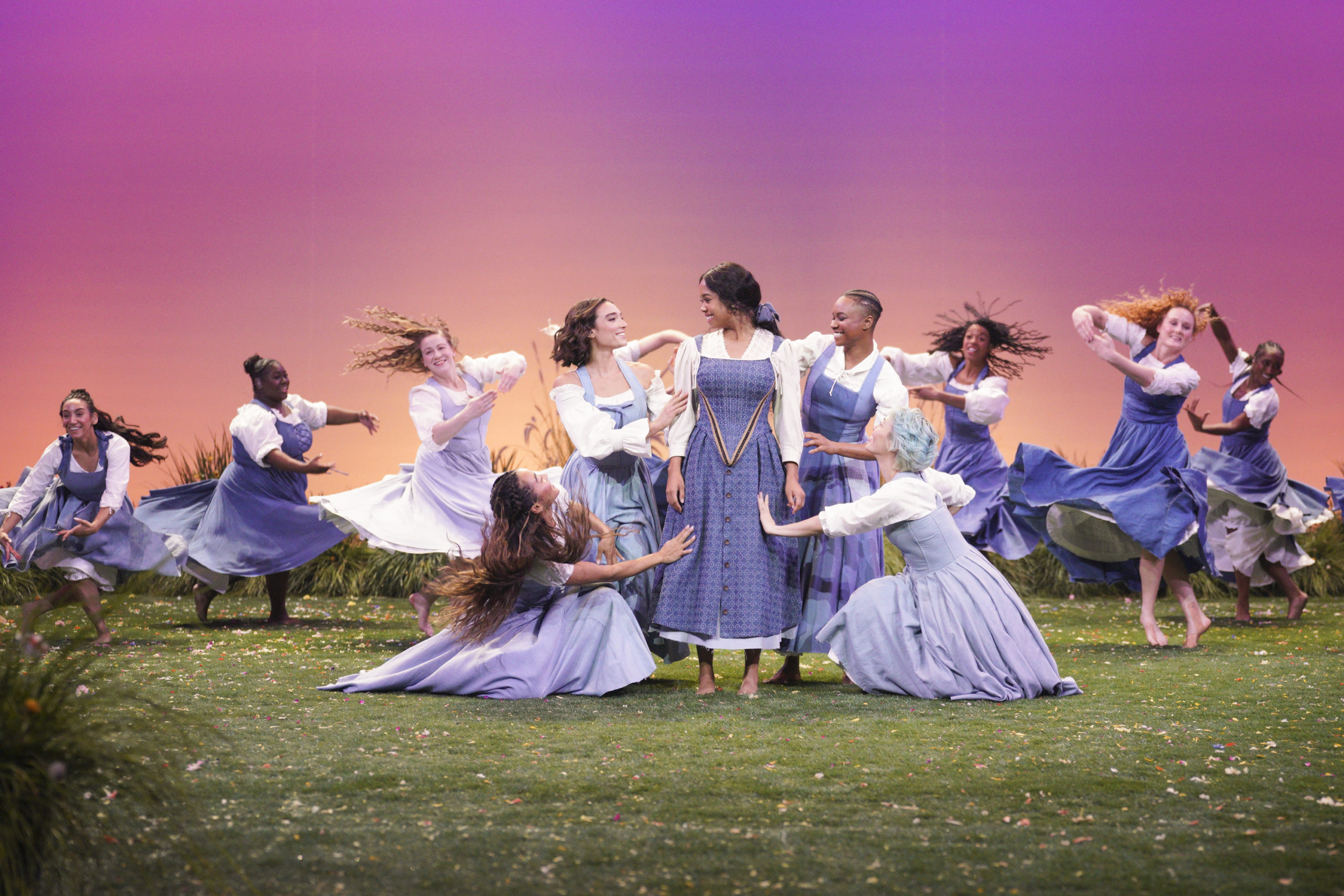 A dancer in a blue dress stands on a green field surrounded by dancers in blue dresses reaching out to her.