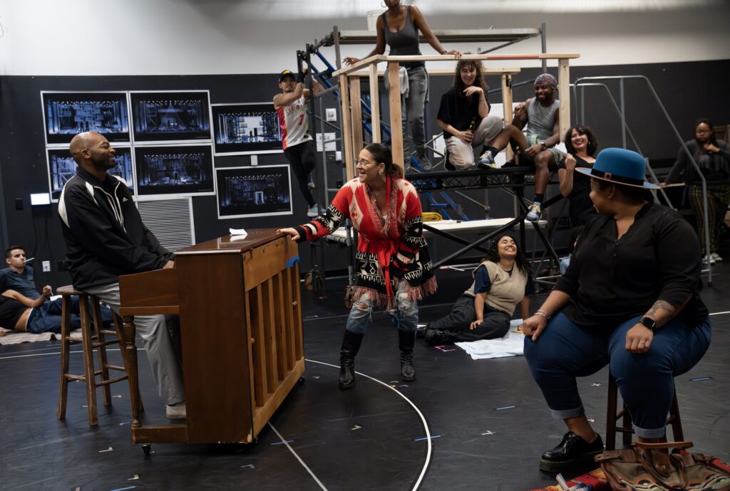 a group of performers surrounding a man at a piano in rehearsal