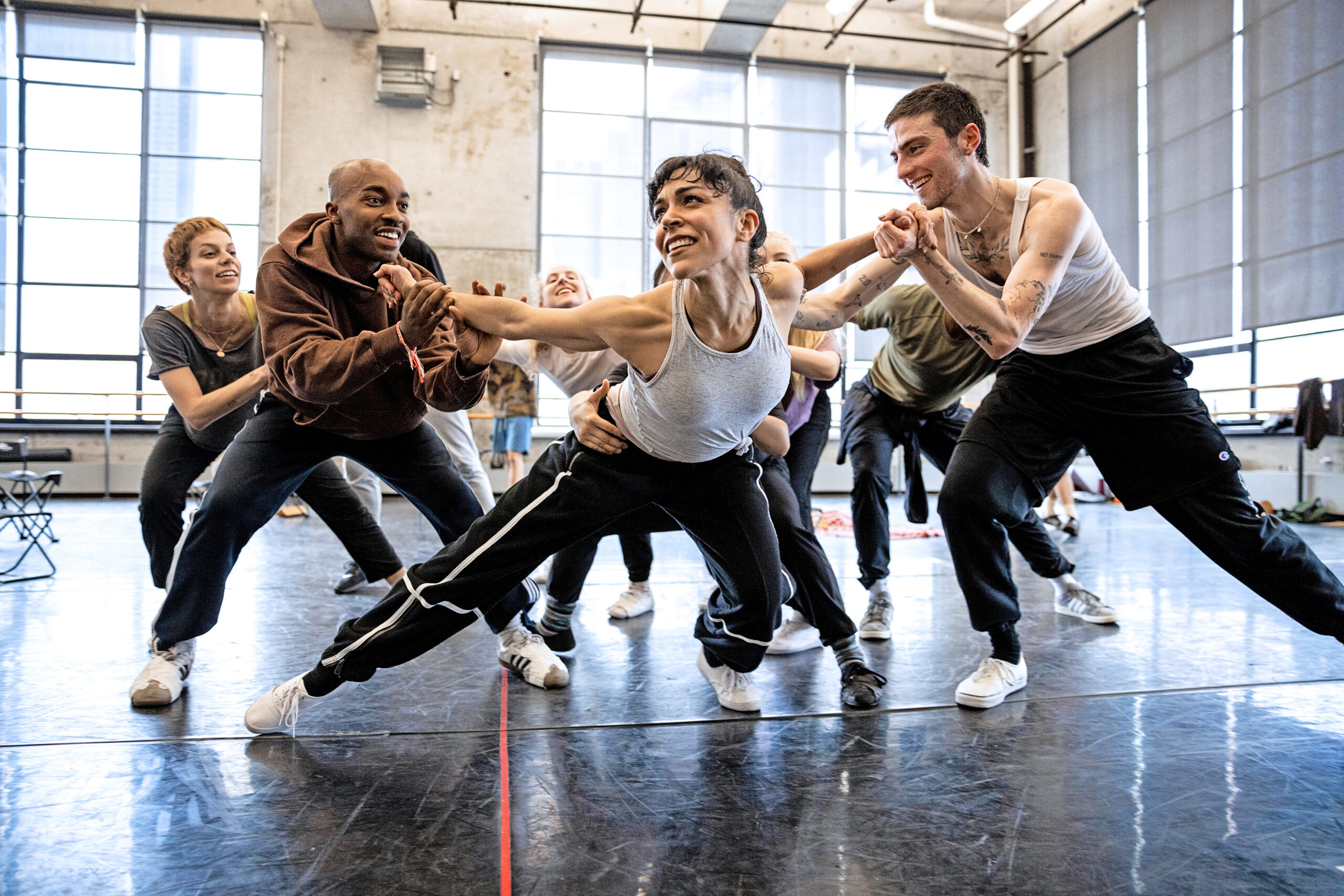 Jeanette Delgado smiles as she lunges to the side and tips forward, looking over her right shoulder. Dancers on either side of her counterbalance her by holding her outstretched arms, while another supports her at the waist from behind.