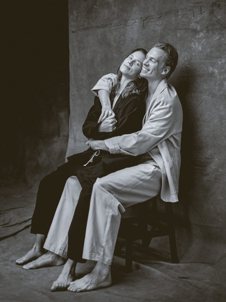 In a black and white image, Imre and Marne van Opstal share a chair. Imre, in black, looks contentedly at the camera, head tipped back against her brother's shoulder and holds one of his hands. Marne, in white, wraps his arms around her, smiling widely.