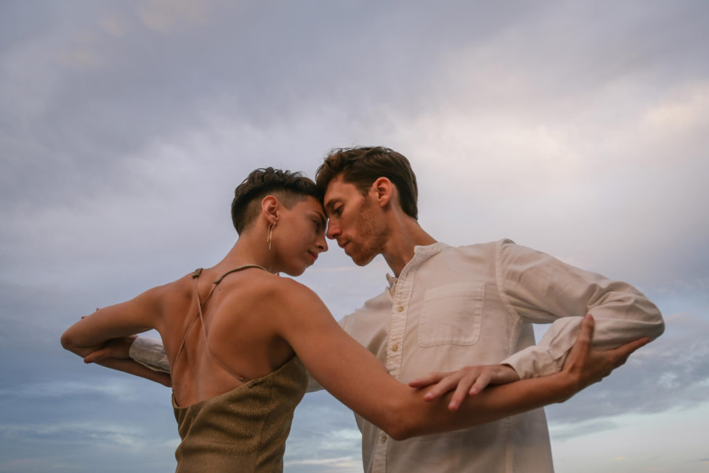 A close-up shot of a male wearing a white, collarless button down shirt and a female dancer in a sparkling gold sleeveless dress face each other against a cloudy blue sky. The woman's hair is short and black with a buzz fade, the man's hair is brown and wavy and he has stubble on his face. Their foreheads are touching, and they are grasping each others' arms right below the elbows.