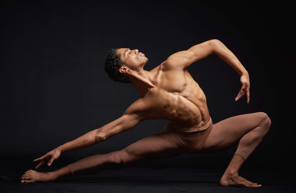 Johnathon Hart lunges, bare chested and barefoot, against a black background. His front arm curves to match the arch of his torso, while his other arm extends parallel to his extended leg. He gazes over his front shoulder proudly.