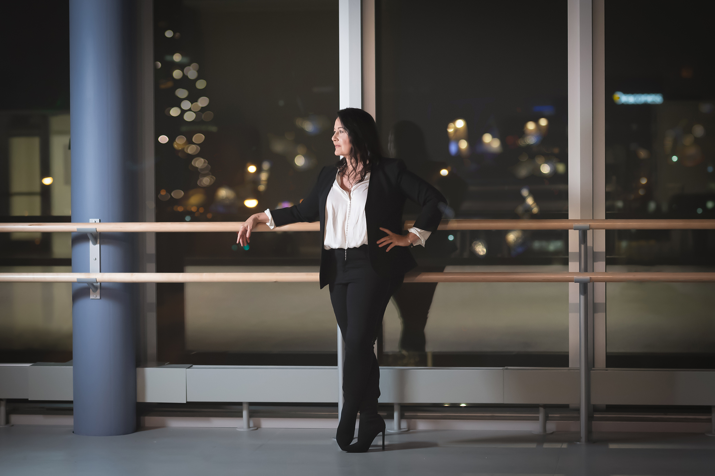 Jodie Gates stands with her back to a ballet barre, floor to ceiling windows behind her showing a city scape at night.