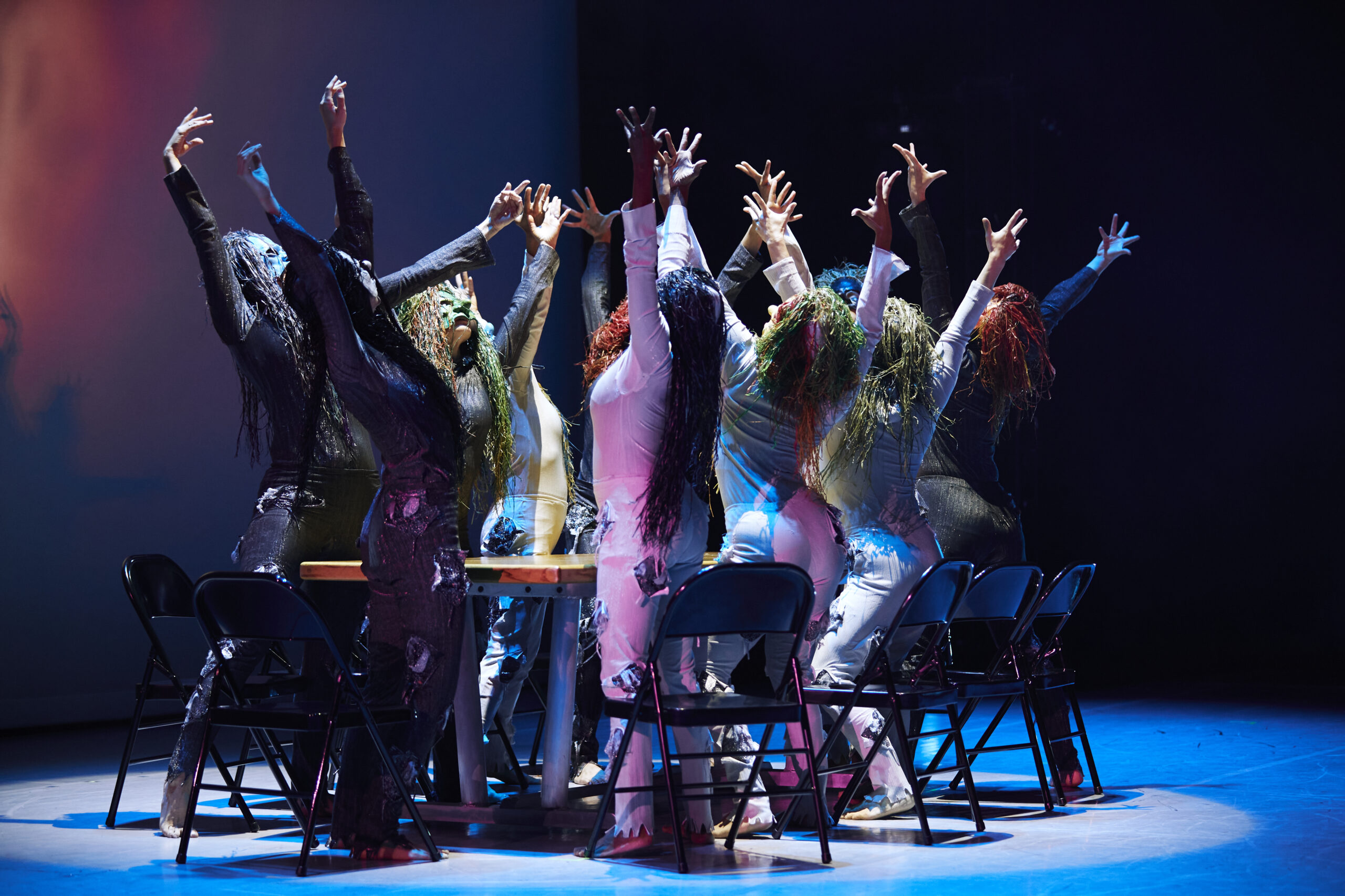 a group of dancers surrounding a table standing out of their chairs with arms lifted overhead