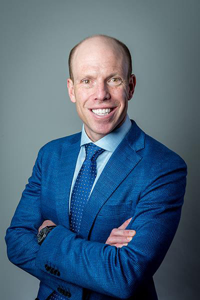 a bald man wearing a blue suit crossing his arms and smiling at the camera