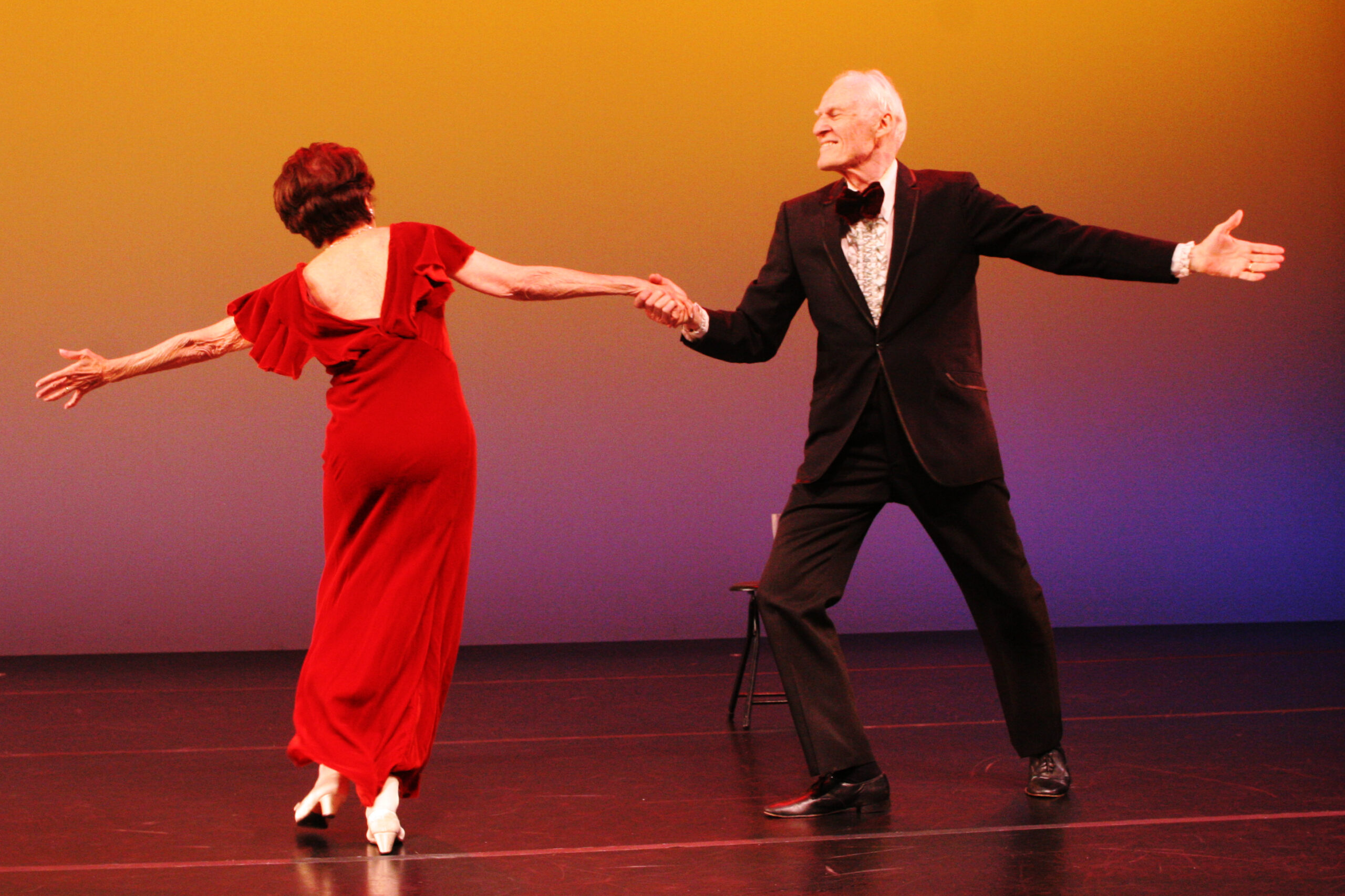 Stuart Hodes, in a black tuxedo, lunges sideways, holding the hand of a woman in a red dress as she leans away from him, balanced on one foot as if ending a turn. They are onstage with an orange backdrop behind them.