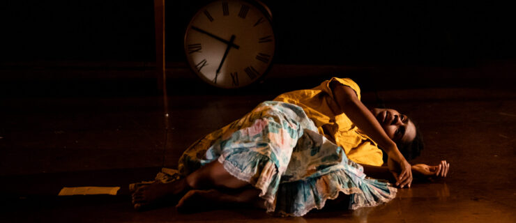 Kayla Farrish is curled up on her side, twisting to stare up at the ceiling. Just behind her, a round clock leaning at an angle against a small table. She wears a bright yellow shirt and a floral patterned skirt.