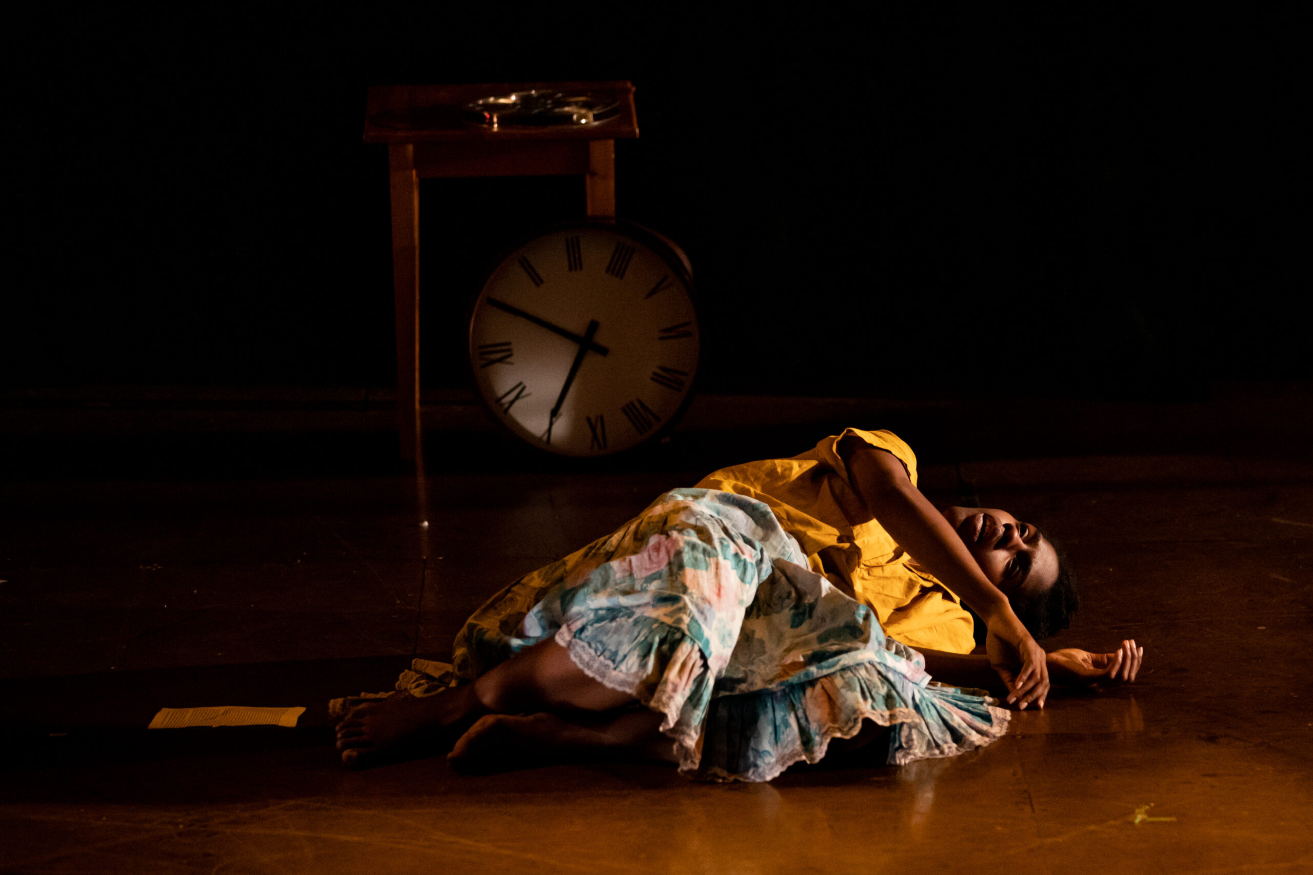 Kayla Farrish is curled up on her side, twisting to stare up at the ceiling. Just behind her, a round clock leaning at an angle against a small table. She wears a bright yellow shirt and a floral patterned skirt.