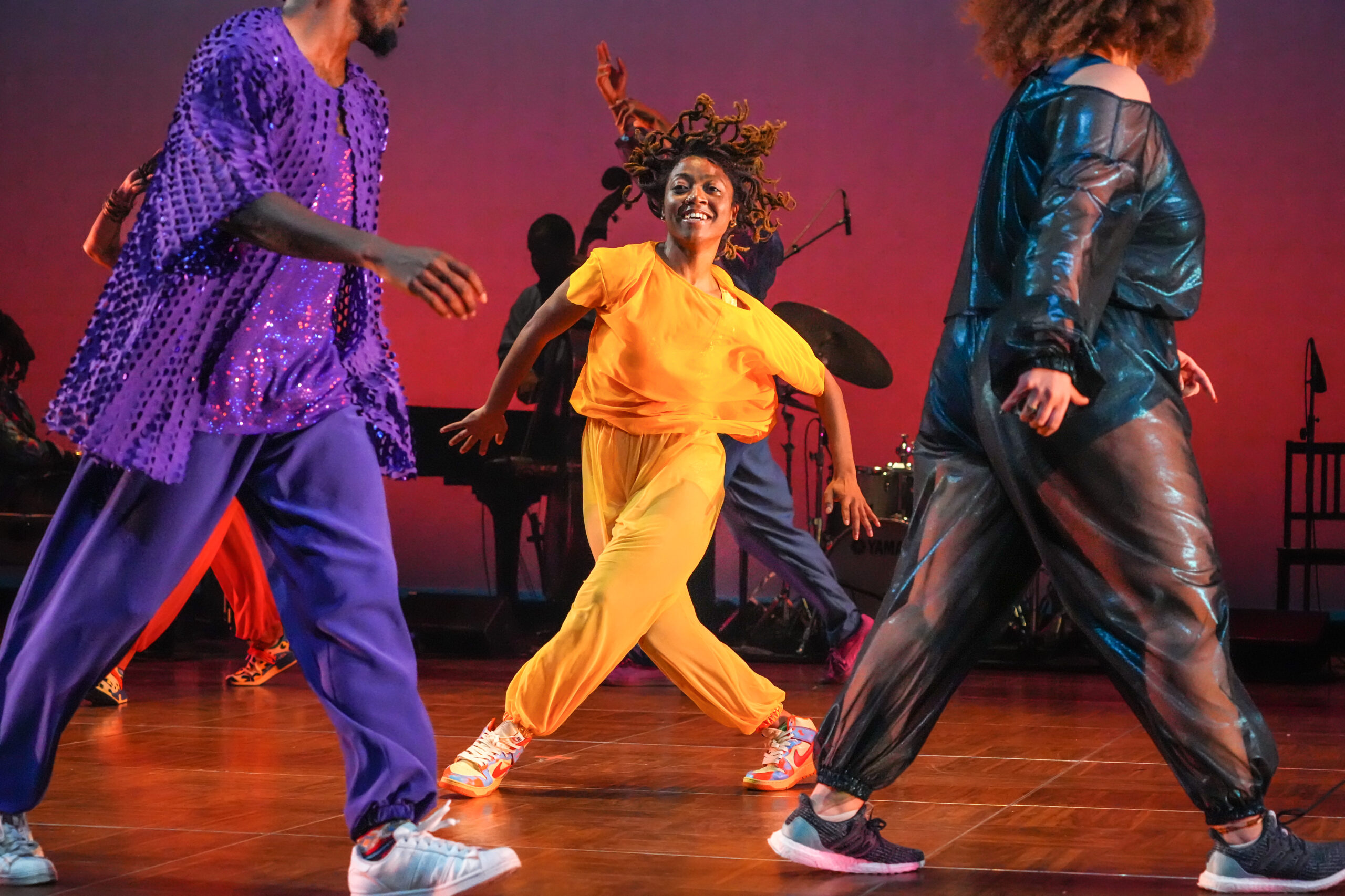A dancer costumed in bright yellow steps into a wide stance, legs crossed. Her arms are raised at the elbows, hair flying up as she begins to turn. In the foreground, a couple of dancers stride around her. Upstage are musicians.