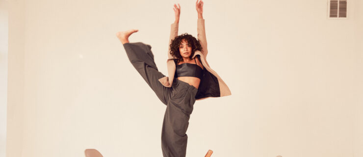a woman wearing brown with a tan coat standing on a wooden chain while extending her leg side with the foot flexed and arms straight up
