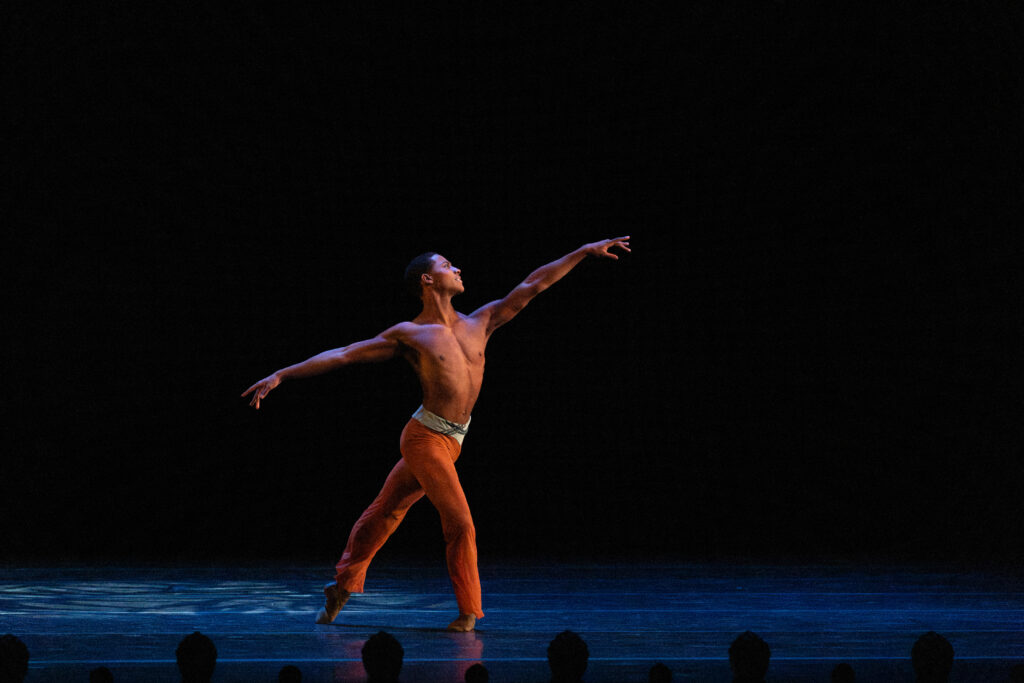 a male dancer wearing orange pants in tendu derriere on stage
