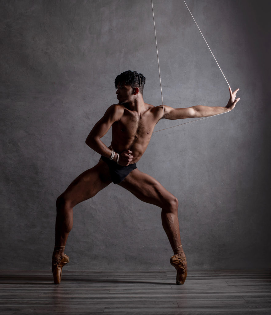 Against a grey backdrop, Maxfield Haynes, wearing tight-fitting shorts and pointe shoes that match their skin tone, poses in a forced arch second position plié on pointe. They look over their right shoulder and shift their ribcage away, opposite arm stretching side with a flexed palm.